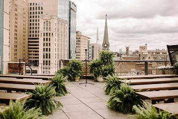 Wedding ceremony setup on The Madison rooftop with brown bench seating flanked by green planters, overlooking downtown Detroit.